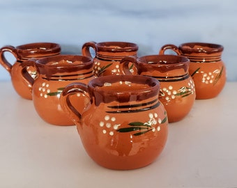 Könnte beinhalten: Satz mit sechs Terrakotta-Bechern mit braunen und grünen Bemalungen. Jeder Becher hat ein weißes Blumenmuster und einen braunen Rand.