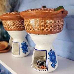 May include: Two terracotta pots with blue and white floral designs, each with a small opening for a heat source. The pots are on white stands with a blue floral design. A small can of solid fuel is visible in the opening of one of the stands.