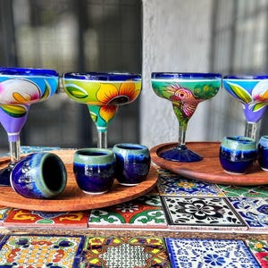 May include: Five hand-painted ceramic margarita glasses with blue rims and colourful floral designs. The glasses are arranged on a wooden tray with four smaller ceramic shot glasses.