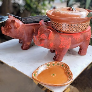 Puede incluir: Dos platos de servir en forma de cerdo de terracota con una olla de terracota con tapa en la parte superior. Los platos en forma de cerdo tienen diseños pintados en rojo y naranja. La olla tiene puntos blancos sobre un fondo marrón. Un plato de terracota en forma de corazón con diseños pintados en verde y blanco está en primer plano.