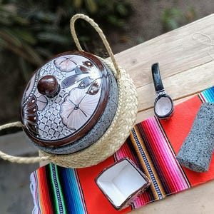 Puede incluir: Un conjunto tradicional mexicano de molcajete y tejolote con una tapa de cerámica pintada a mano. El conjunto se muestra en una estera de paja tejida con una manta mexicana colorida debajo. El conjunto incluye un pequeño tazón de cerámica y una cuchara de madera.