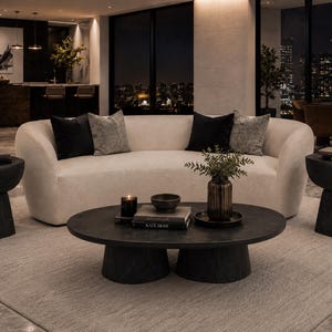May include: A curved cream-colored sofa with two black and two gray throw pillows, flanked by two black pedestal side tables. A matching black oval coffee table sits in front, topped with books, a small bowl, a candle, and a vase with greenery.