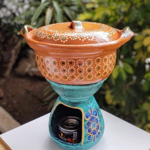 May include: A terracotta pot with a lid and a blue and gold base. The pot has a white and green dotted design. The base has a blue floral design. A small can of solid fuel is visible inside the base.