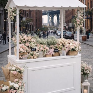 Puede incluir: Un puesto de flores blanco en una calle adoquinada, lleno de varios ramos y arreglos de flores rosas y blancas. El puesto tiene un techo festoneado y está decorado con detalles florales. El fondo presenta una calle de la ciudad y un puente.
