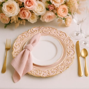 May include: A formal table setting with a pink and gold decorative charger plate, white plate, and pink napkin. Gold flatware includes a fork, knife, and spoon. A bouquet of peach and cream roses and greenery is in the background.