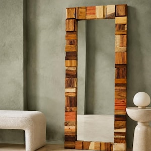 Puede incluir: Un espejo de suelo rectangular y alto con un marco construido a partir de numerosos bloques de madera pequeños y apilados en varios tonos de marrón, bronceado y naranja rojizo. La superficie del espejo es clara y refleja el espacio.