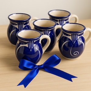 Puede incluir: Un juego de cinco tazas de cerámica azul cobalto con asas blancas y diseños decorativos. Una cinta de raso azul en primer plano. Las tazas tienen una forma tradicional.