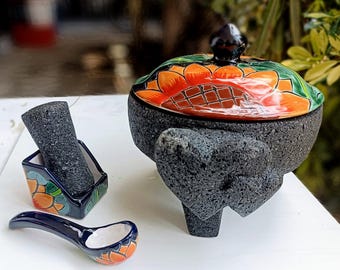 Puede incluir: Un juego tradicional mexicano de molcajete y tejolote con una tapa de cerámica pintada a mano. El molcajete está hecho de roca volcánica y presenta un diseño floral en tonos naranja, verde y azul. El juego también incluye una pequeña cuchara de cerámica.
