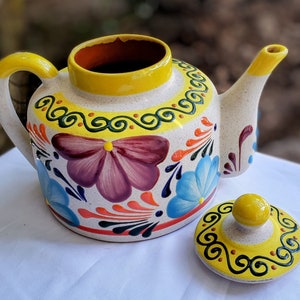 Puede incluir: Una tetera de cerámica blanca con tapa y asa amarillas, decorada con diseños florales coloridos y un borde negro y amarillo. La tetera tiene una tapa a juego con un pomo amarillo.