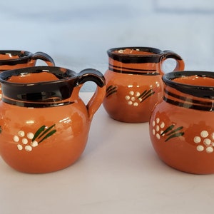 May include: Set of four orange ceramic mugs with black and green painted designs. Each mug has a white floral pattern and a black rim.