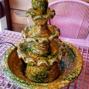 May include: A three-tiered ceramic fountain with a green and brown marbled design. Water flows from the top tier down to the bottom basin.