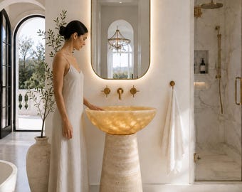 May include: A pedestal sink featuring a translucent, glowing amber-coloured stone basin atop a tapered, textured beige stone base. The sink is paired with gold-toned wall-mounted taps and positioned beneath an arched, backlit mirror in a bathroom.