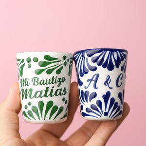 Puede incluir: Dos pequeños vasos de chupito de cerámica blanca con diseños pintados a mano. Uno tiene patrones florales verdes y el texto "Mi Bautizo Matias". El otro tiene patrones florales azules y las letras "A & C."