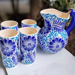 Puede incluir: Un juego de cuatro vasos para beber de cerámica azul y blanca y una jarra a juego con un diseño floral. La jarra tiene un asa azul.