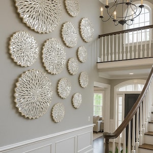 Puede incluir: Una colección de piezas decorativas de arte mural de varios tamaños. Las piezas circulares tienen un diseño texturizado de color plateado con un borde dorado. El arte se exhibe en una pared gris claro en la entrada de una casa.