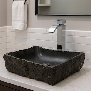 May include: A dark grey stone basin with a textured surface, rectangular in shape, sits on a white countertop. A chrome tap is above the sink. A white towel hangs on a black towel ring.