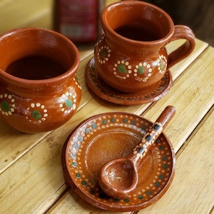 Puede incluir: Dos tazas de barro marrón con diseños pintados en verde y blanco, una con un platillo a juego, y una cuchara de barro marrón en un platillo a juego.