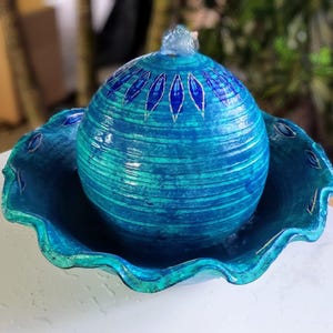 May include: A blue ceramic fountain with a wavy rim and a round, textured top. The fountain has a blue and white design with a pattern of leaves.