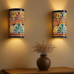 May include: Two cylindrical wall sconces with colourful floral patterns in orange, yellow, and teal. The sconces are mounted on a beige wall, with a wooden table and a vase of dried flowers in the foreground. The sconces have a dark blue trim.