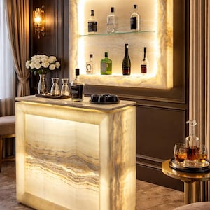 Puede incluir: Una barra de bar doméstica de ónix retroiluminada con una encimera de mármol, que presenta una estantería con botellas de licor. La barra tiene un brillo cálido y acogedor, complementado con una pequeña mesa auxiliar con una jarra y vasos.