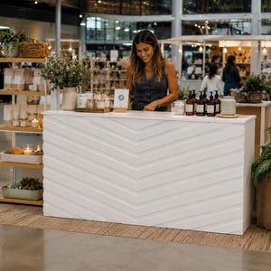 Puede incluir: Un mostrador de exhibición rectangular blanco con un patrón de espiga en el panel frontal. El mostrador está ubicado en un espacio comercial, acompañado por una estantería de madera alta llena de varios productos y una planta grande en maceta.
