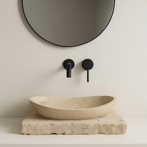 May include: A contemporary bathroom arrangement with a beige stone basin on a matching stone plinth. Above, a black tap and a circular black mirror are mounted on a white wall, creating a minimalist aesthetic.