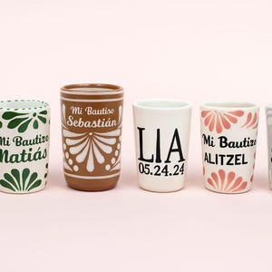 Puede incluir: Seis vasos de chupito de cerámica con varios diseños y texto. Los vasos presentan colores azul, verde, marrón, blanco y gris. Algunos vasos tienen diseños florales, mientras que otros muestran nombres y fechas. Los vasos de chupito están sobre una superficie rosa claro.