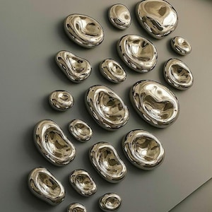 May include: A cluster of polished, silver-toned, organic-shaped wall decorations. The pieces are of varying sizes and are mounted on a grey wall, creating a modern, abstract design. The reflective surfaces catch the light, adding depth and interest.
