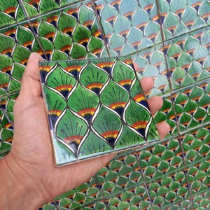 Puede incluir: Un solo azulejo de cerámica verde, azul, amarillo y naranja con un patrón repetitivo de hojas estilizadas. El azulejo se está sosteniendo en una mano.