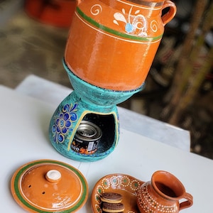 May include: A terracotta clay pot with a green and white floral design sits on a teal blue stand with a floral design. The stand has a small opening for a heat source. A small terracotta clay cup with a brown and white dot design sits on a terracotta clay plate with a green and white dot design. The plate has four chocolate sandwich cookies on it.