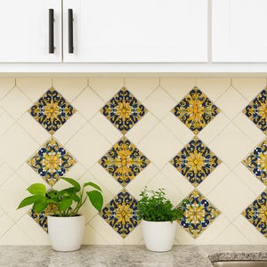 Puede incluir: Salpicadero de cocina color crema con azulejos en forma de diamante con un diseño floral amarillo y azul. Dos pequeñas plantas en macetas se encuentran en la encimera. Armarios blancos con tiradores negros encima.