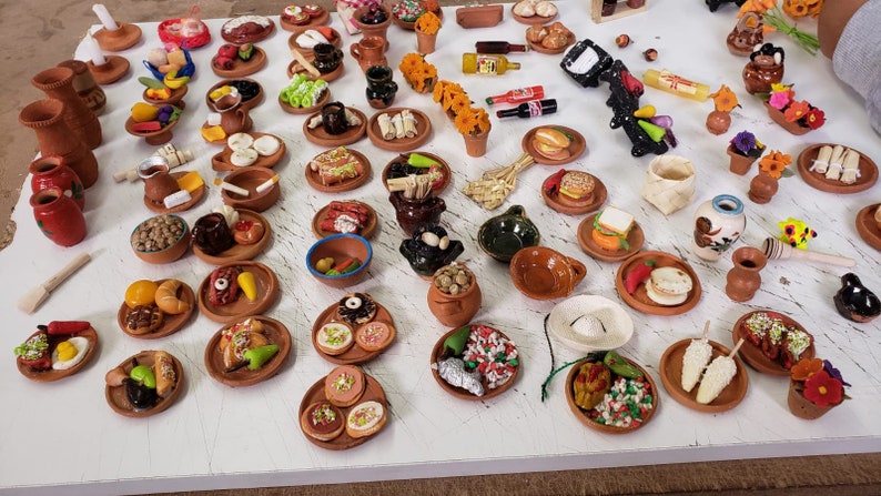 Puede incluir: Una colecci&oacute;n de alimentos en miniatura de arcilla sobre peque&ntilde;os platos de arcilla. Los alimentos incluyen tacos, tamales, churros y otros platos mexicanos. Los platos est&aacute;n dispuestos sobre una superficie blanca.