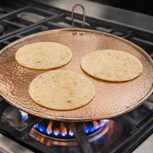 Puede incluir: Un calentador de tortillas de color cobre con tres tortillas encima, calentado por una estufa de gas. El calentador redondo tiene un asa y se encuentra directamente sobre las llamas azules. El electrodoméstico de cocina está diseñado para cocinar tortillas.