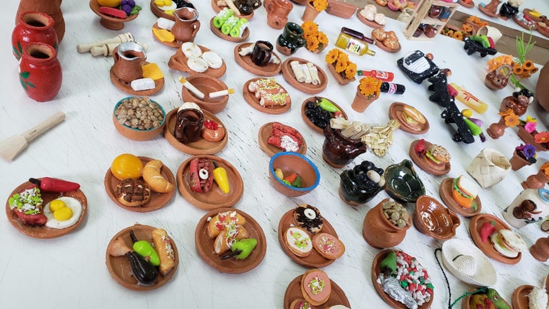 Puede incluir: Una colecci&oacute;n de platos de arcilla en miniatura llenos de varios alimentos, incluyendo huevos, pimientos, pan y fruta. Los platos est&aacute;n dispuestos sobre una superficie blanca.