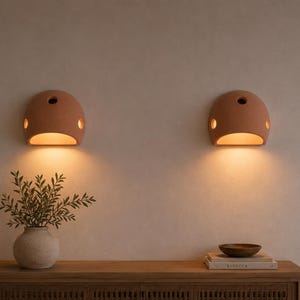 Puede incluir: Dos apliques de pared semicirculares de terracota montados uno al lado del otro, cada uno con una abertura central y dos recortes laterales más pequeños que emiten un brillo cálido hacia abajo. Acabado mate y color arcilla natural.