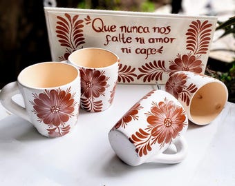 Puede incluir: Cuatro tazas de café de cerámica blanca con diseños florales marrones. Las tazas están sobre una superficie blanca con un letrero que dice "Que nunca nos falte ni amor ni cafe".