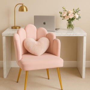 Puede incluir: Una silla de escritorio rosa con patas doradas y una almohada en forma de corazón. Un escritorio con tapa de mármol sostiene una computadora portátil, una lámpara de escritorio dorada y un jarrón con rosas rosas. La silla tiene respaldo festoneado.
