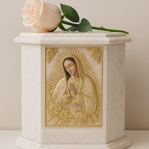 May include: A white marble urn with a gold relief of a religious figure, adorned with roses. A light pink rose rests on top. The urn has an octagonal shape and is likely used for memorial purposes.