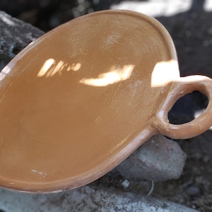 Puede incluir: Un plato ovalado para servir de terracota con un solo asa. El plato no está esmaltado y tiene un aspecto rústico.