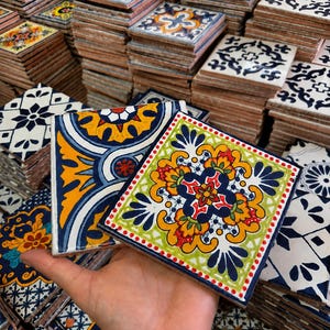 Puede incluir: Dos azulejos de cerámica cuadrados con complejos patrones pintados a mano. Un azulejo muestra un audaz motivo floral en amarillo, azul y blanco, mientras que el otro presenta un diseño simétrico vibrante en verde, amarillo, rojo y azul.