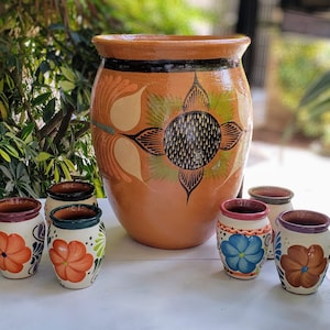 May include: Six colorful ceramic mugs with floral designs. The mugs are arranged in a semi-circle around a larger, brown ceramic pitcher with a floral design.