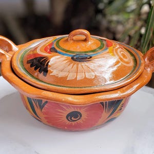 May include: A terracotta casserole dish with a lid, featuring colorful floral designs in shades of red, orange, yellow, green, blue, and white. The dish has a scalloped edge and two handles.