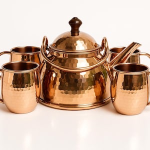 May include: A copper tea kettle with a hammered finish, a dark wooden handle, and a set of four matching copper mugs. The kettle has a large handle and a spout. The mugs have a handle on the side.