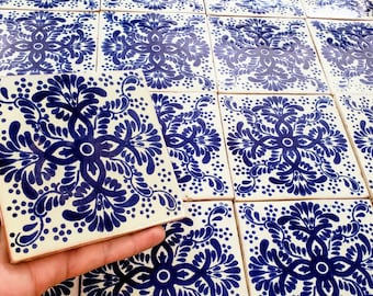 May include: A close-up of a hand holding a single white ceramic tile with a blue floral pattern. The tile is surrounded by other similar tiles, creating a repeating pattern.
