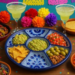 Op de afbeelding: Een levendige afbeelding van een Mexicaans feest met een blauw-witte verdeelde serveerschaal gevuld met guacamole, salsa, olijven en amandelen. Tortillachips zijn ook inbegrepen. Twee margarita-cocktails en kleurrijke papieren bloemen maken de feestelijke scène compleet.
