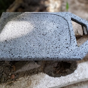 Puede incluir: Una tabla de servir gris moteada con un asa. La tabla tiene forma rectangular con esquinas redondeadas y una zona empotrada. El asa tiene forma geométrica. La tabla está hecha de un material similar a la piedra.