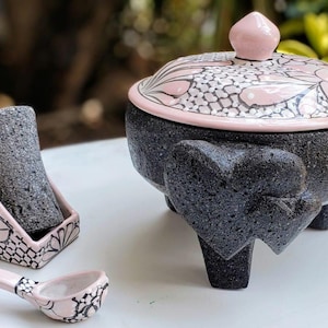 May include: A black volcanic stone mortar and pestle set with a pink and white floral design. The mortar has three legs and the pestle is in a matching stand.