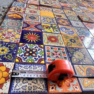 May include: A collection of colourful, hand-painted ceramic tiles with various floral and geometric patterns. The tiles are arranged in a grid pattern and measure approximately 4 inches by 4 inches. A tape measure is placed on top of the tiles, showing the measurement in inches.