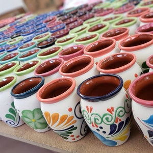 Puede incluir: Una colección de pequeñas tazas de cerámica pintadas a mano con diseños florales coloridos. Las tazas tienen una base blanca con un borde marrón y están dispuestas en filas sobre una superficie de madera.