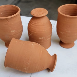 Puede incluir: Cuatro tazas de arcilla de terracota, dos con una base más ancha y dos con una base más estrecha. Las tazas son todas de un tono marrón similar y tienen una superficie rugosa y texturizada.
